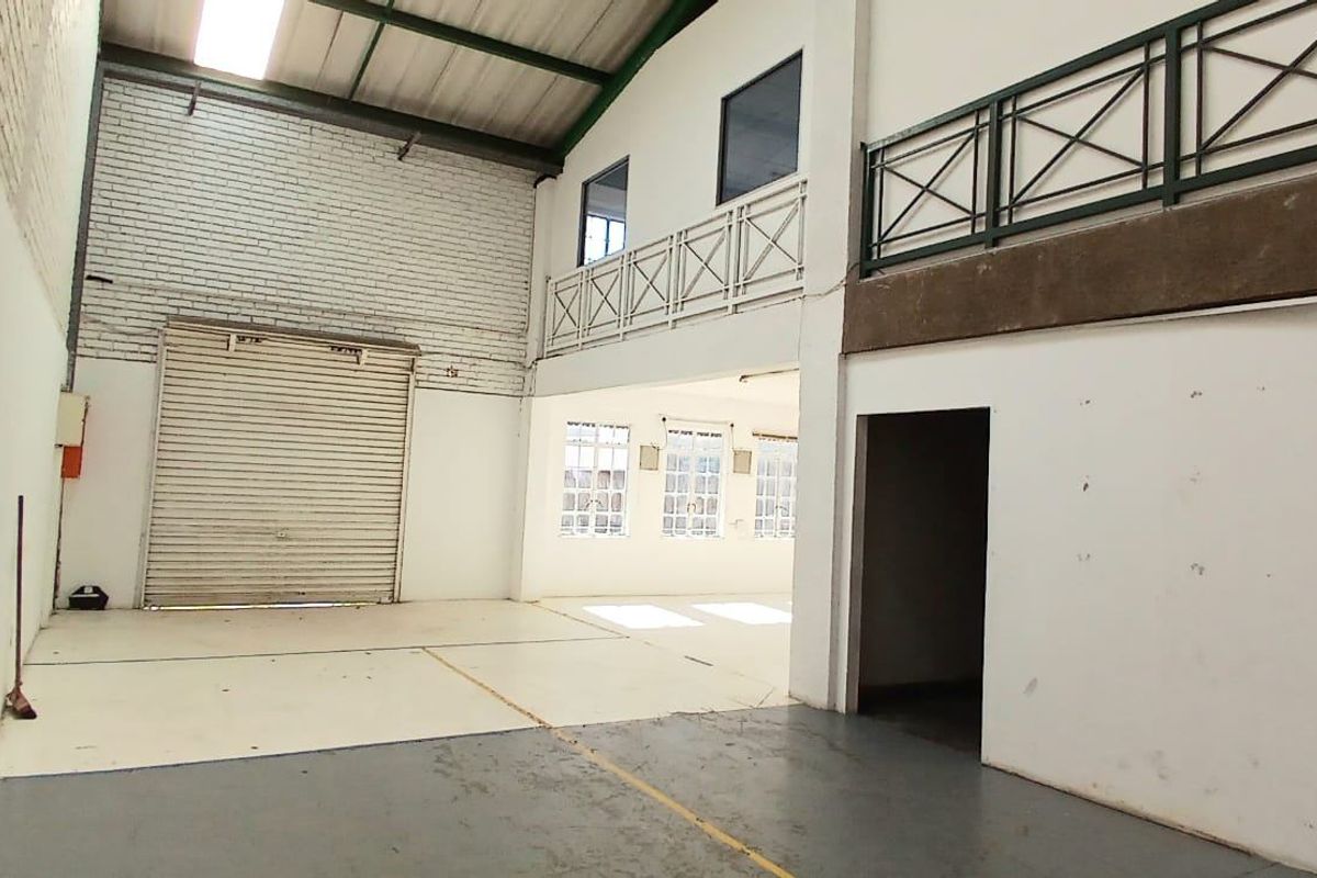 Spacious industrial utility area featuring a high-clearance roller shutter door, mezzanine level with metal railing, and exposed roof structure with natural skylight illumination.