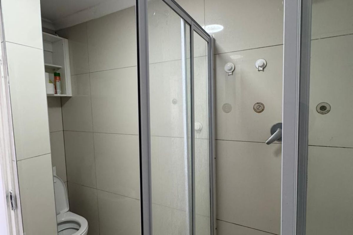 A functional bathroom featuring a glass shower enclosure, tiled walls and floors, and a standard toilet with built-in storage shelving.