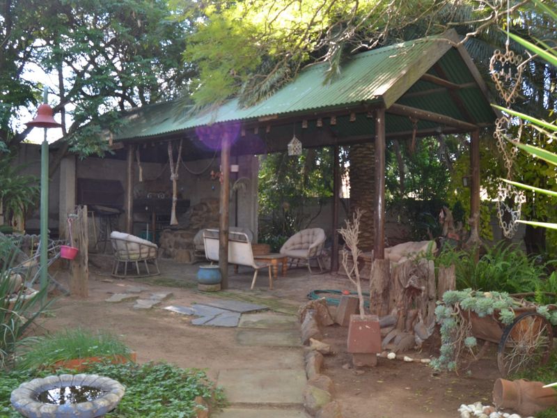 A rustic outdoor patio area featuring a corrugated metal roof, timber support posts, and a built-in braai, set within a garden environment.