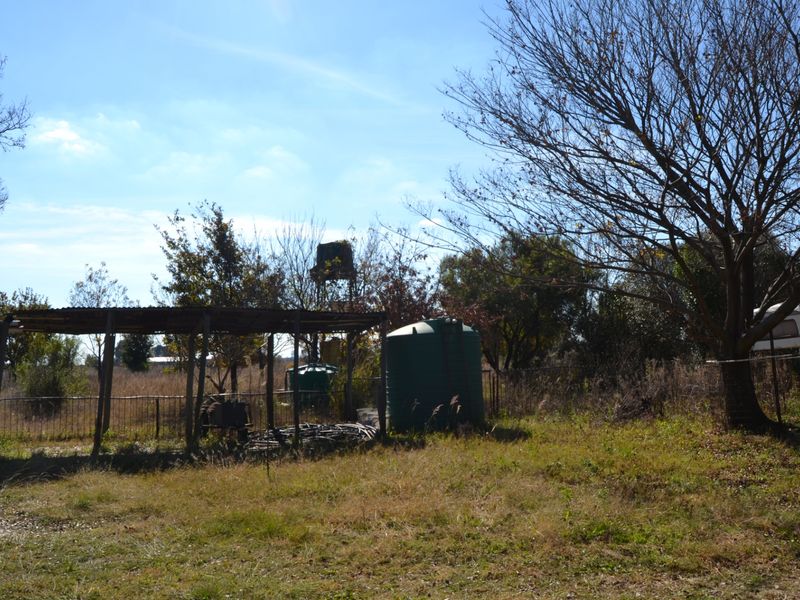 A vacant residential plot featuring an open-sided shelter, water storage tanks, and mature trees on a grassy landscape.