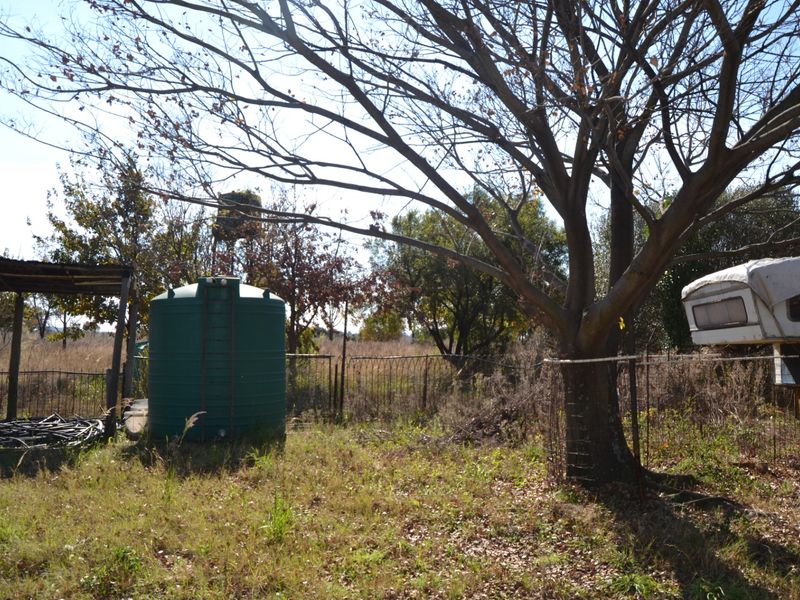 A view of the residential land featuring a large water tank, mature trees, and perimeter wire fencing on an overgrown plot.