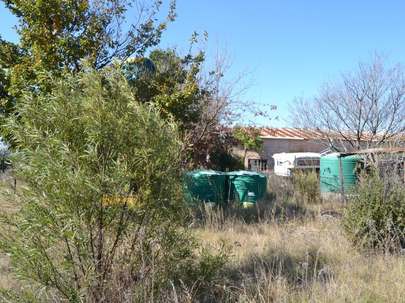 Rear view of the property showing a structure with a rusted corrugated metal roof and several green water storage tanks amidst overgrown vegetation.