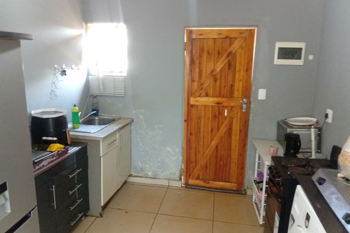 A functional kitchen featuring tiled flooring, dark and light cabinetry, a stainless steel sink, and a wooden exterior door with visible wall surface damage.