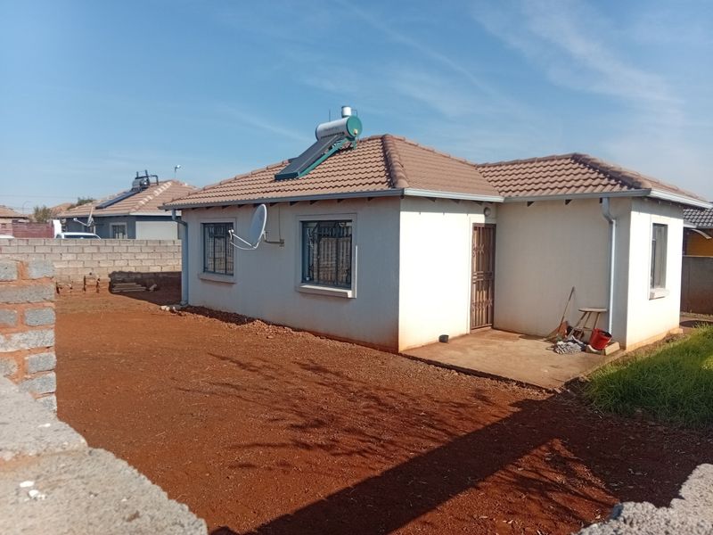 Rear view of a single-story house featuring a tiled roof, solar water heater, security bars, and a concrete patio area with an unlandscaped yard.