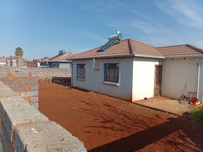 Rear view of a single-story house featuring a tiled roof, solar water heater, security bars on windows, and an unlandscaped yard with boundary walls.