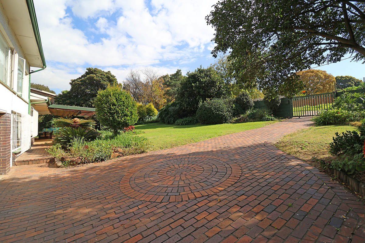 A spacious rear garden featuring a circular brick paved driveway, established landscaping, mature trees, and a covered patio area.