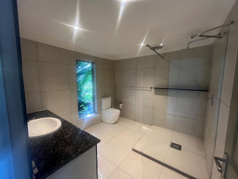 Modern bathroom featuring a walk-in shower with glass screen, granite-topped vanity, and neutral floor-to-ceiling tiling with natural light.