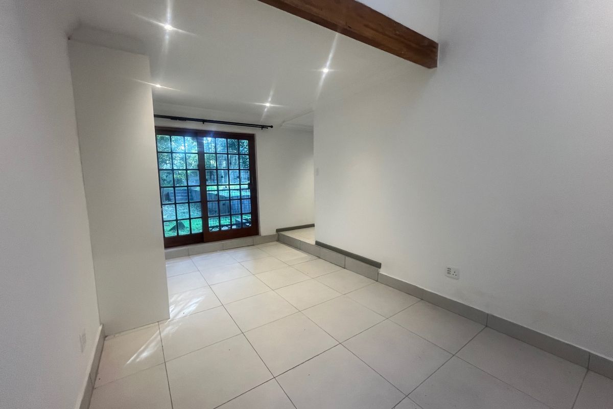 A bright, empty living area featuring tiled floors, recessed lighting, an exposed timber beam, and sliding glass doors leading to the garden.