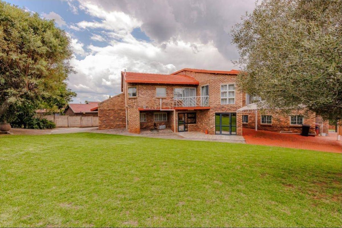 Rear view of a double-story brick house featuring a tiled roof, balcony, paved patio area, and a spacious garden.