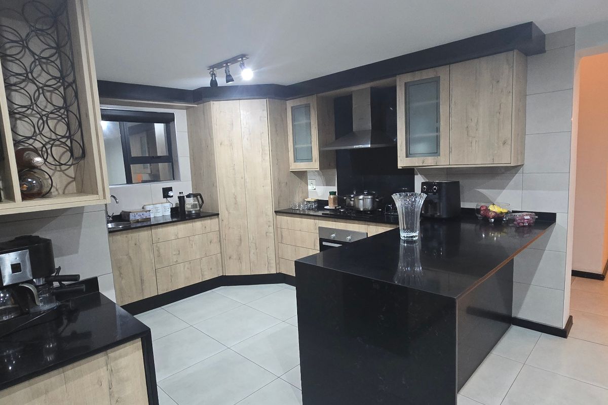 Modern kitchen featuring light wood-tone cabinetry, contrasting black granite countertops, a central island, and integrated appliances with sleek tiled flooring.