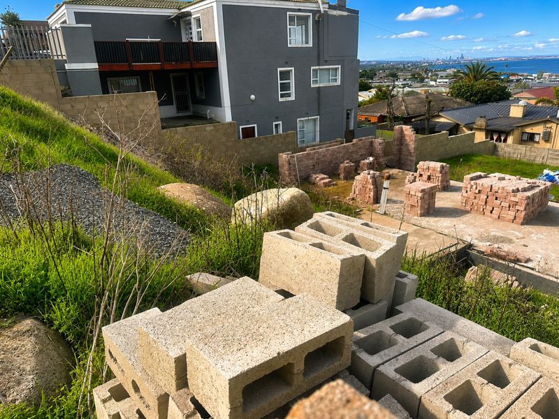 A sloped residential plot featuring ongoing construction work, including stacked concrete blocks and partial brick wall foundations with distant coastal views.