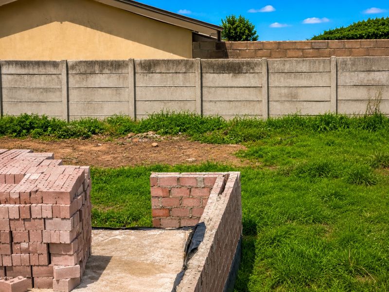 A vacant residential plot featuring a concrete boundary wall and ongoing construction work with stacked bricks on a concrete foundation.