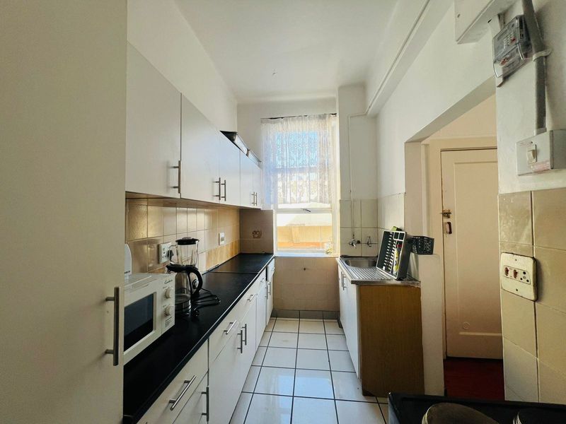 A functional galley-style kitchen featuring white cabinetry, dark laminate countertops, tiled flooring, and a window providing natural light.