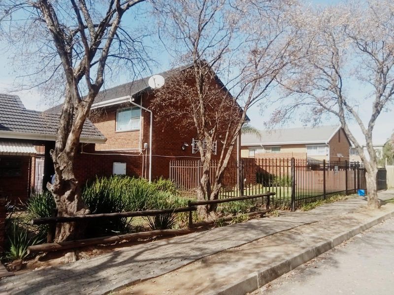 Street view of a brick apartment complex featuring a tiled roof, metal security fencing, and established trees along the sidewalk.