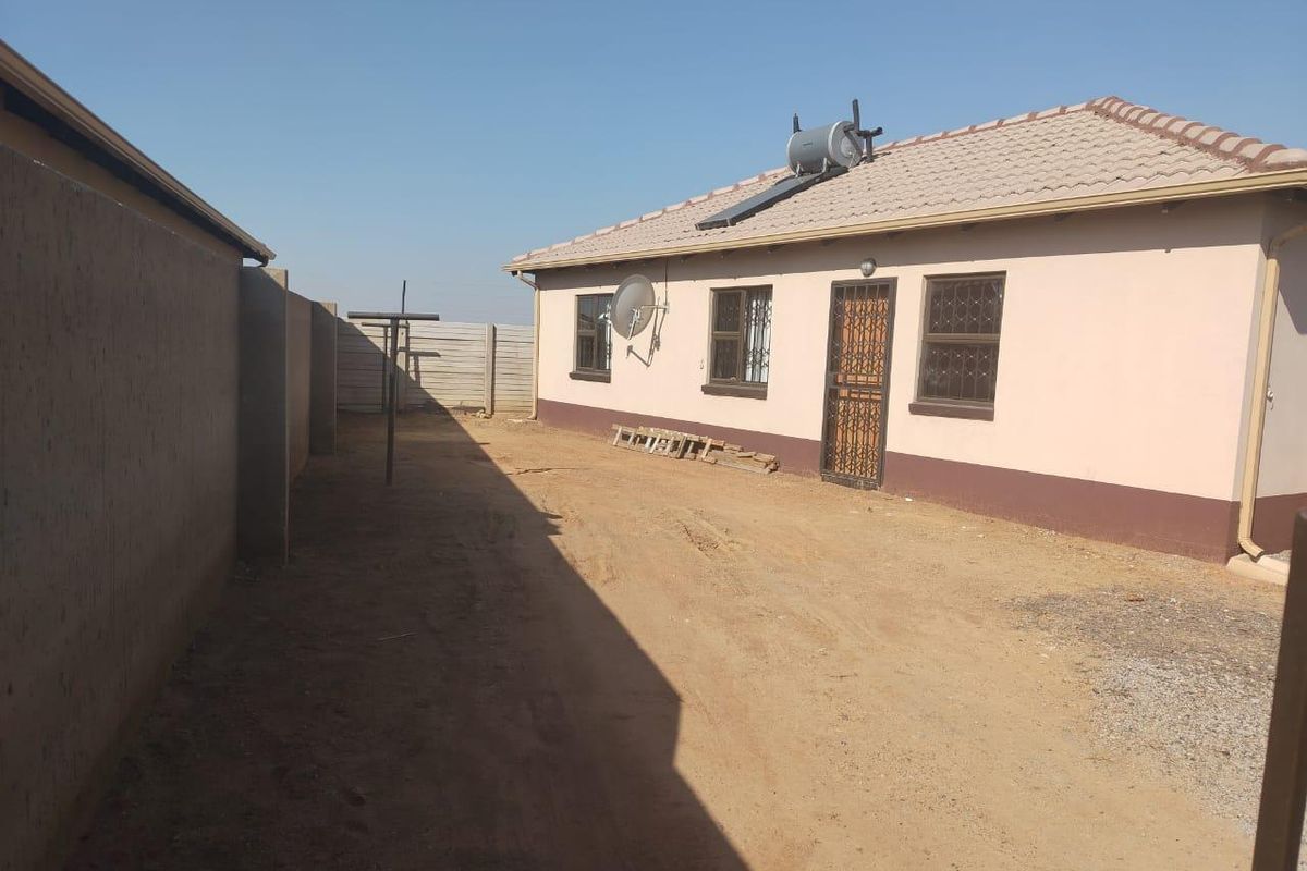 Rear exterior view of the house featuring a solar water heater, security gates, and a large unpaved yard area enclosed by precast concrete walls.