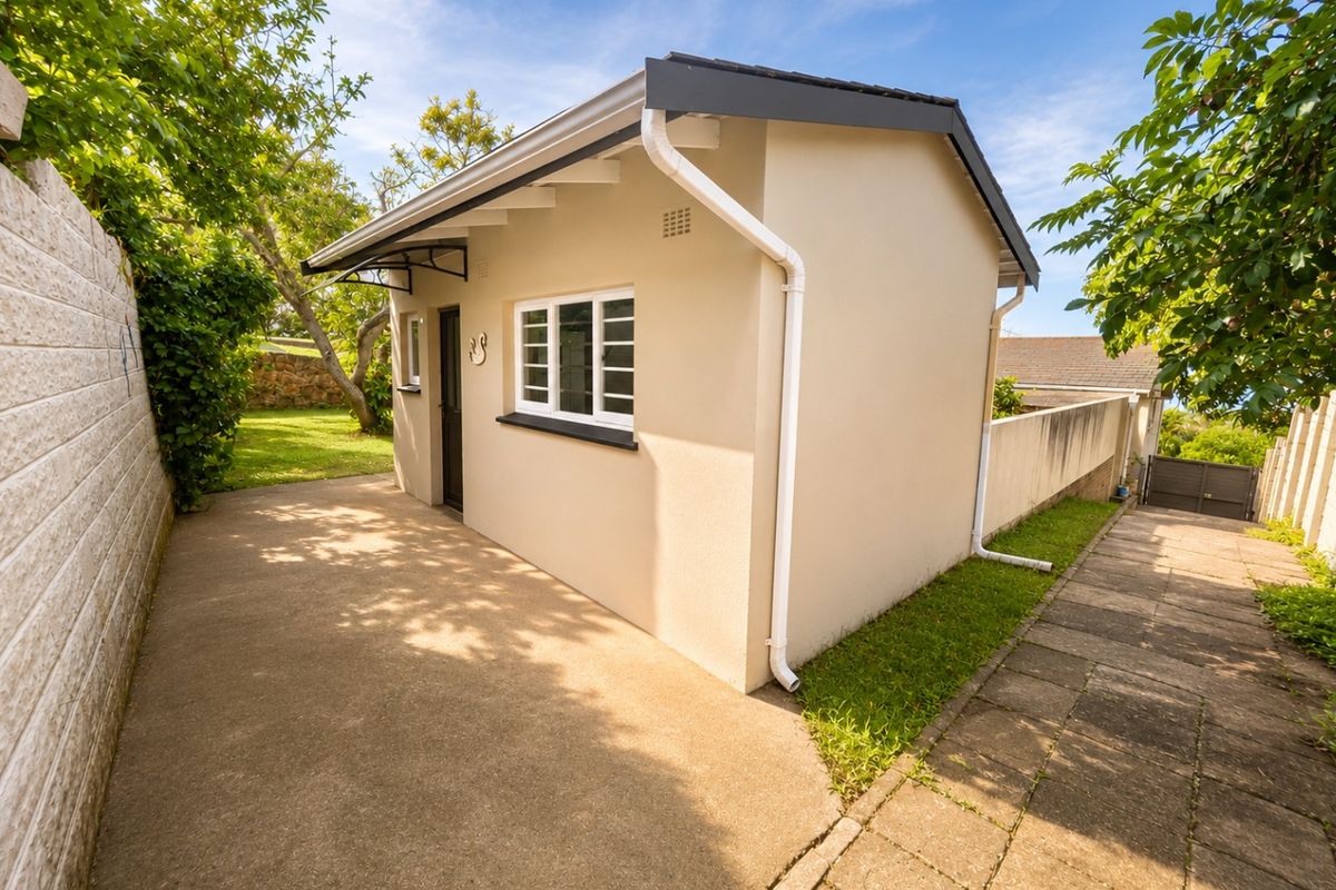 A well-maintained exterior view of a small outbuilding featuring concrete paving, clean render, and functional guttering systems set within a landscaped garden area.