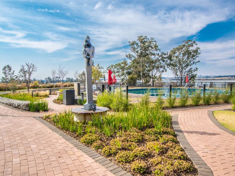 A well-maintained outdoor pool area featuring paved walkways, landscaped garden beds, and a central water fountain within a secure residential complex.