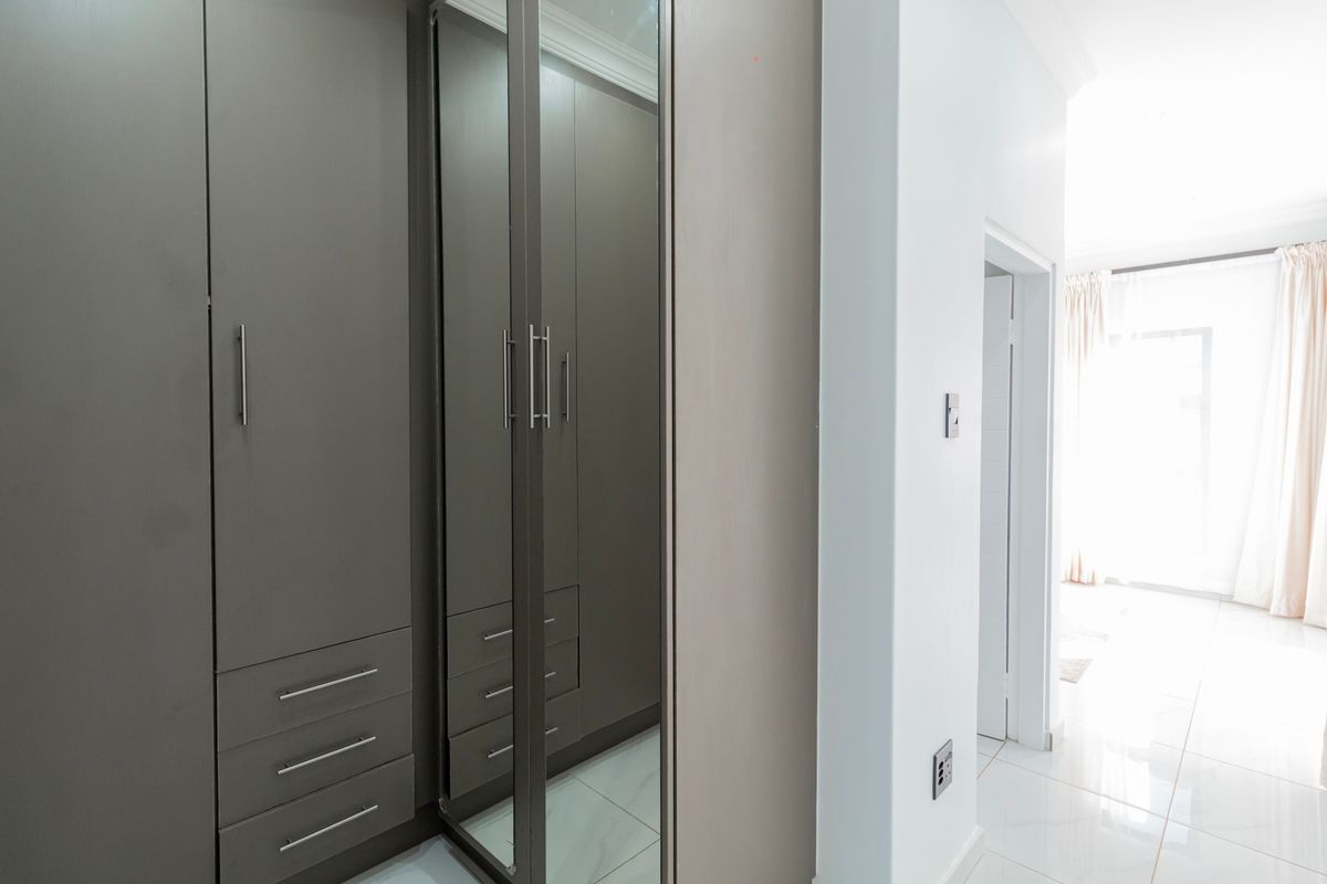 A bright bedroom featuring modern built-in cupboards with mirrored doors and clean tiled flooring, leading into an adjacent living space.