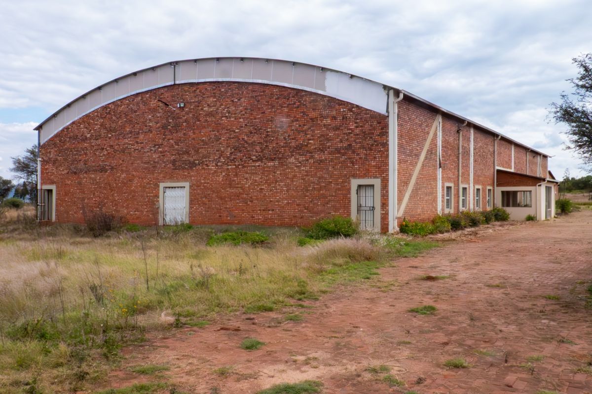 Large industrial-style farm building featuring a curved metal roof and red brick exterior, situated on a rural property with a paved access area.