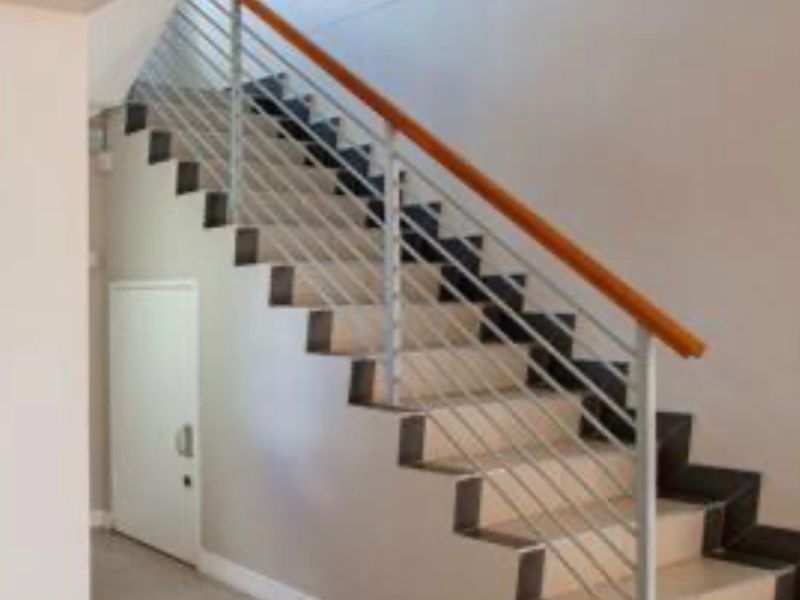 A clean, modern hallway featuring a staircase with a metal balustrade, wooden handrail, and neutral tiled flooring.