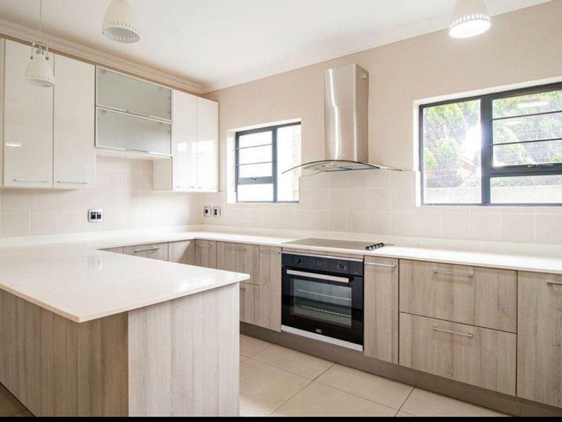 Modern kitchen featuring light wood-grain cabinetry, stone countertops, built-in oven and hob, stainless steel extractor, and neutral tiled finishes.