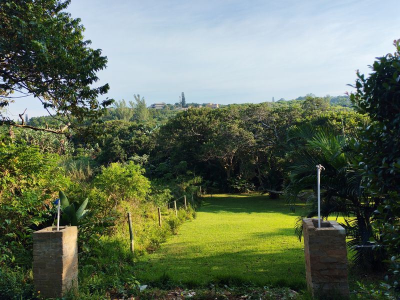 A well-maintained residential garden plot featuring brick entrance pillars, wire fencing, and lush green lawn surrounded by mature trees and vegetation.