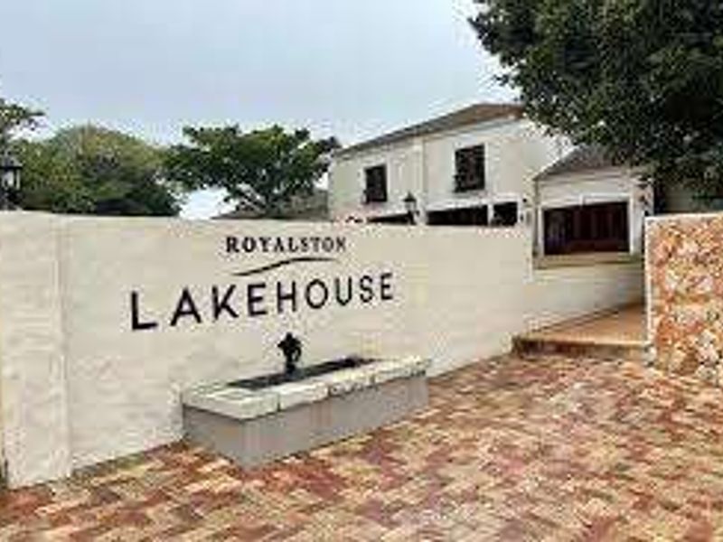 Entrance view of the Royalston Lakehouse featuring a decorative boundary wall, stone cladding accents, brick paved driveway, and a built-in water feature.