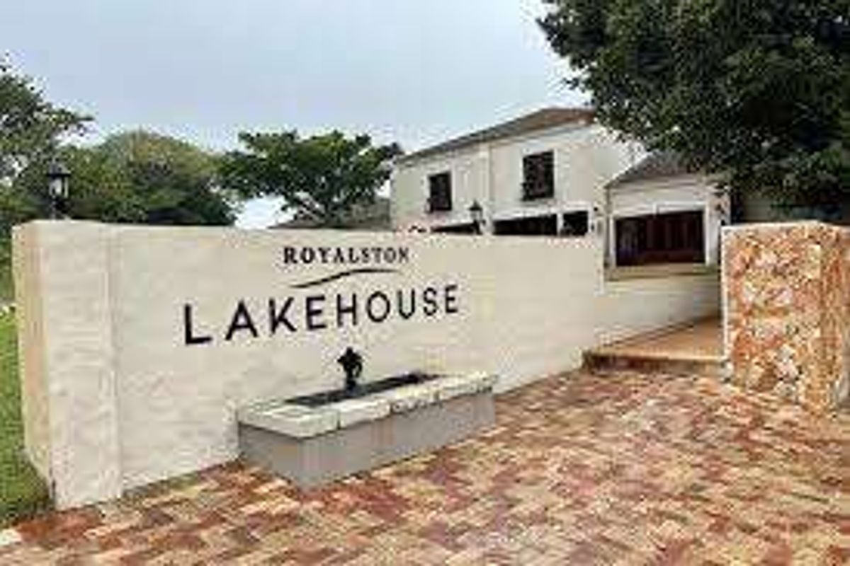 Entrance view of the Royalston Lakehouse featuring a decorative boundary wall, stone cladding accents, brick paved driveway, and a built-in water feature.
