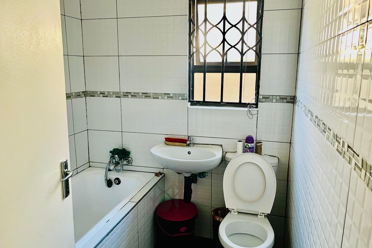 A functional bathroom featuring a bathtub, pedestal basin, and toilet, finished with tiled walls and fitted with security bars on the window.