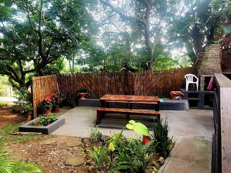 Outdoor patio area featuring a built-in brick braai, concrete flooring, and timber balustrades, set within a lush, tree-lined garden environment.