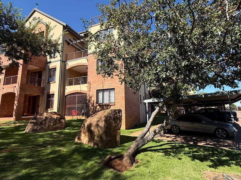 Exterior view of a multi-story apartment building featuring brick facade, private balconies, and an adjacent carport area set within a landscaped garden.