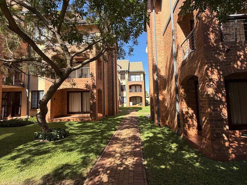 A view of the apartment complex exterior featuring brick facades, private balconies, and a well-maintained garden with a paved walkway.