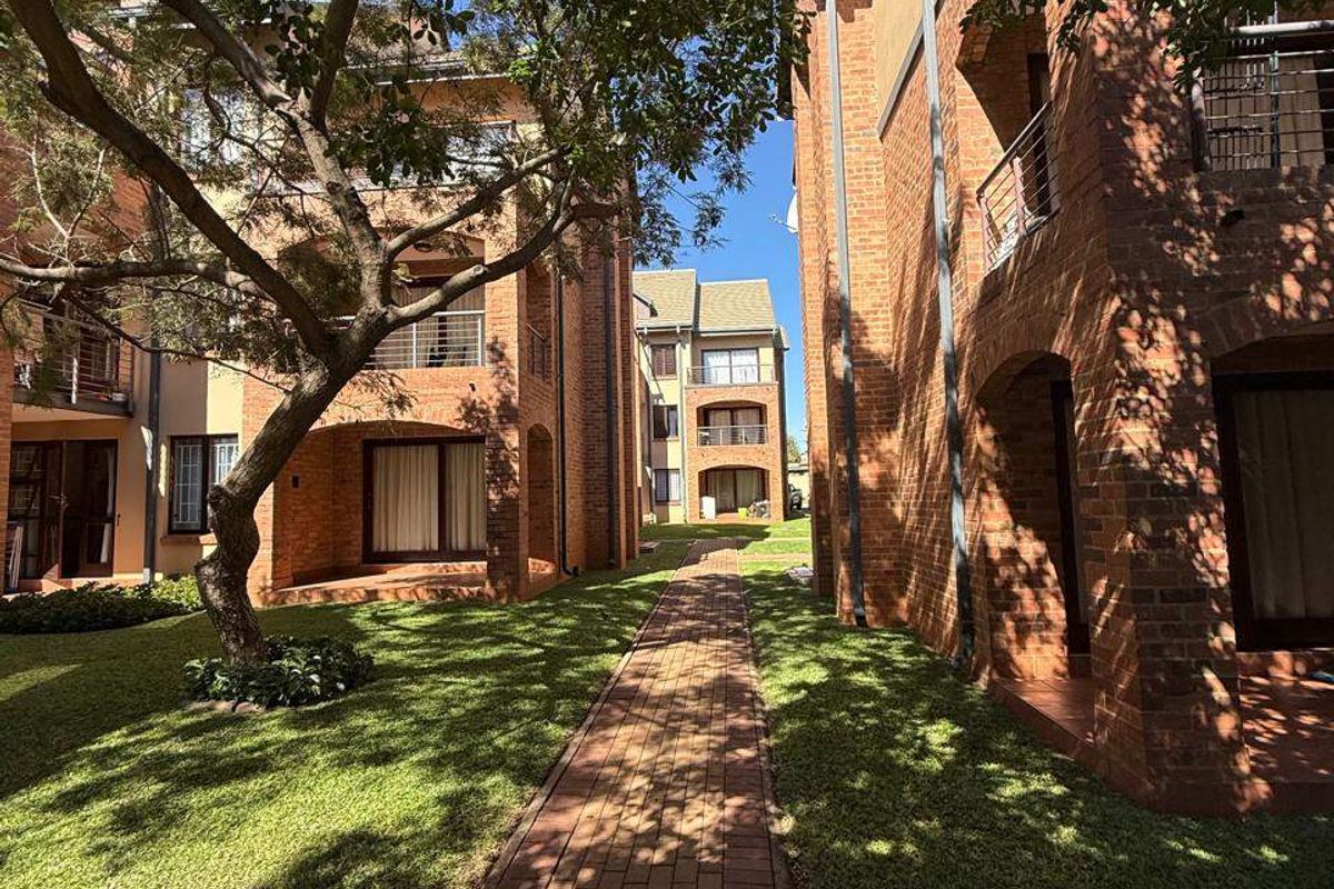 A view of the apartment complex exterior featuring brick facades, private balconies, and a well-maintained garden with a paved walkway.