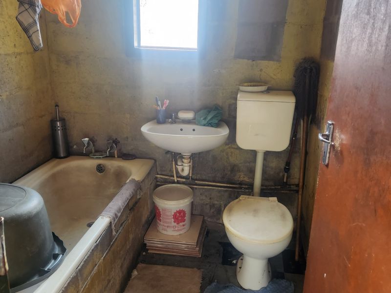 A small, dated bathroom featuring a bathtub, wall-mounted basin, and toilet, with unfinished brick walls requiring significant renovation and cosmetic updates.