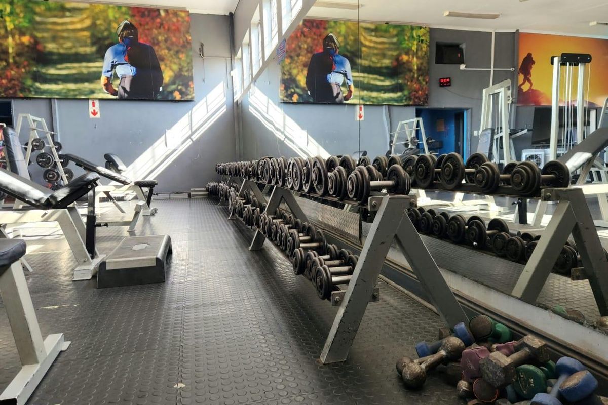 A commercial gym facility featuring rubber flooring, a large wall-mounted mirror, and overhead lighting, equipped with various weight training stations.