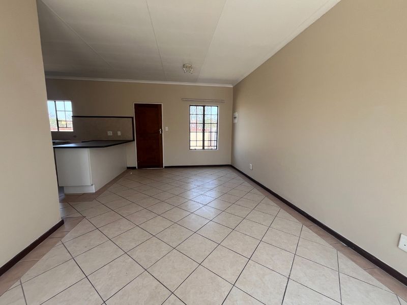 An open-plan living and kitchen area featuring neutral tiled flooring, a breakfast bar, and natural light through security-fitted windows.