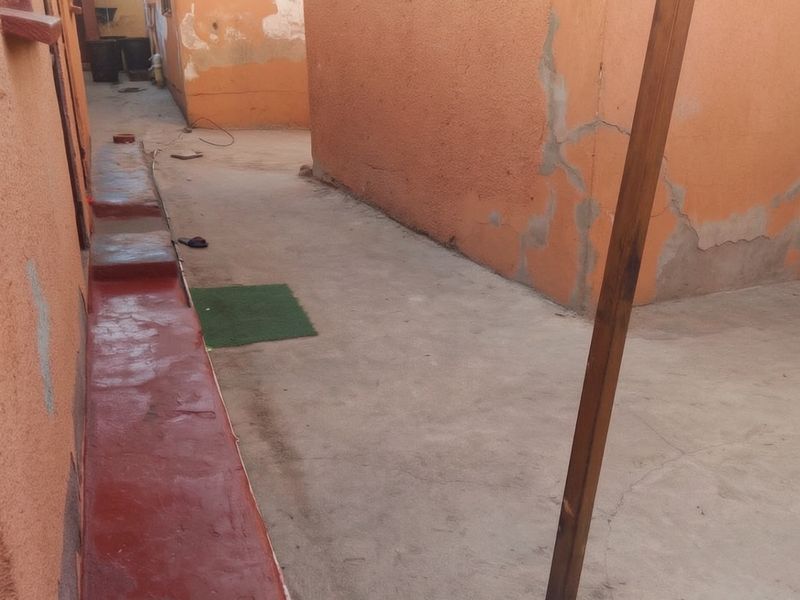A narrow rear exterior walkway featuring concrete paving and painted walls showing signs of wear and plaster cracking.