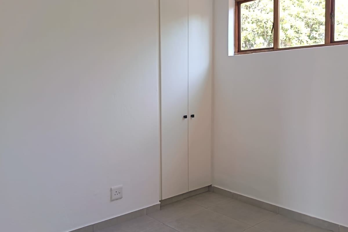 A clean, empty bedroom featuring neutral tiled flooring, a built-in cupboard, and a timber-framed window providing natural light.