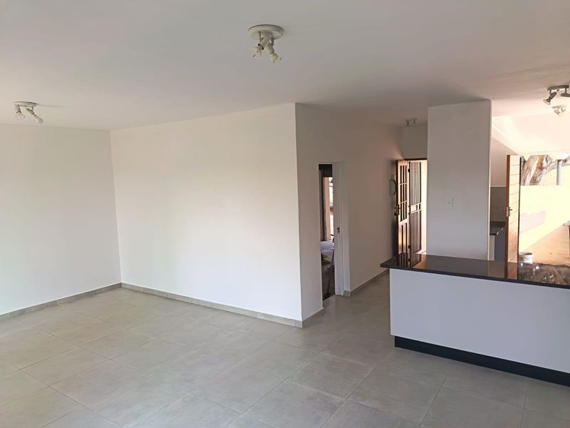 Spacious open-plan living area featuring neutral tiled flooring, a modern granite kitchen breakfast bar, and direct access to the exterior via a security gate.