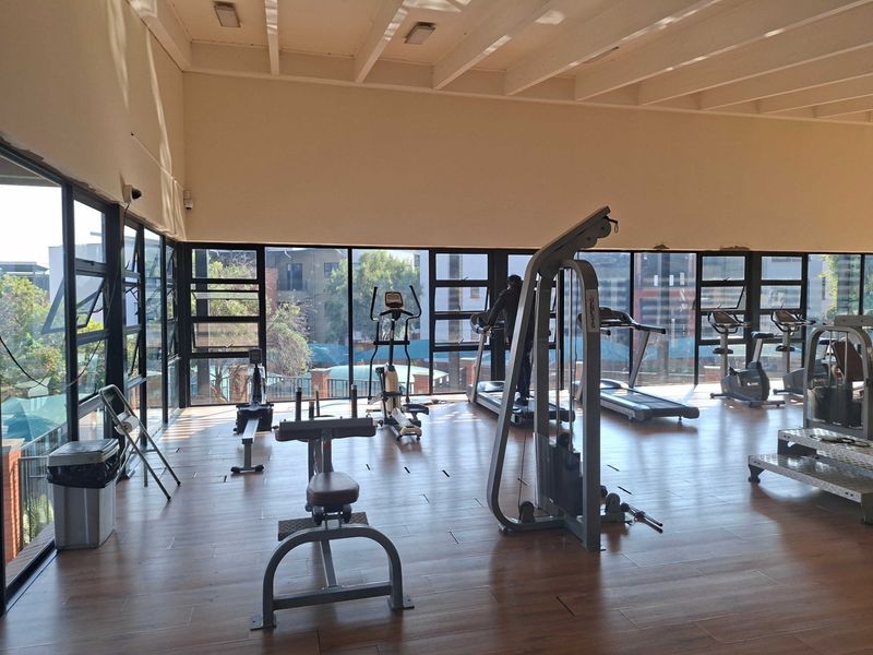 A spacious apartment gym featuring large floor-to-ceiling windows, laminate flooring, and an exposed beam ceiling, providing a bright and functional workout environment.