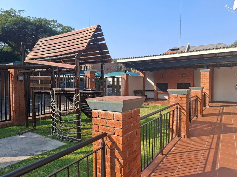 Outdoor communal area featuring a tiled patio, built-in braai facilities, and a wooden playground structure set within a landscaped garden space.