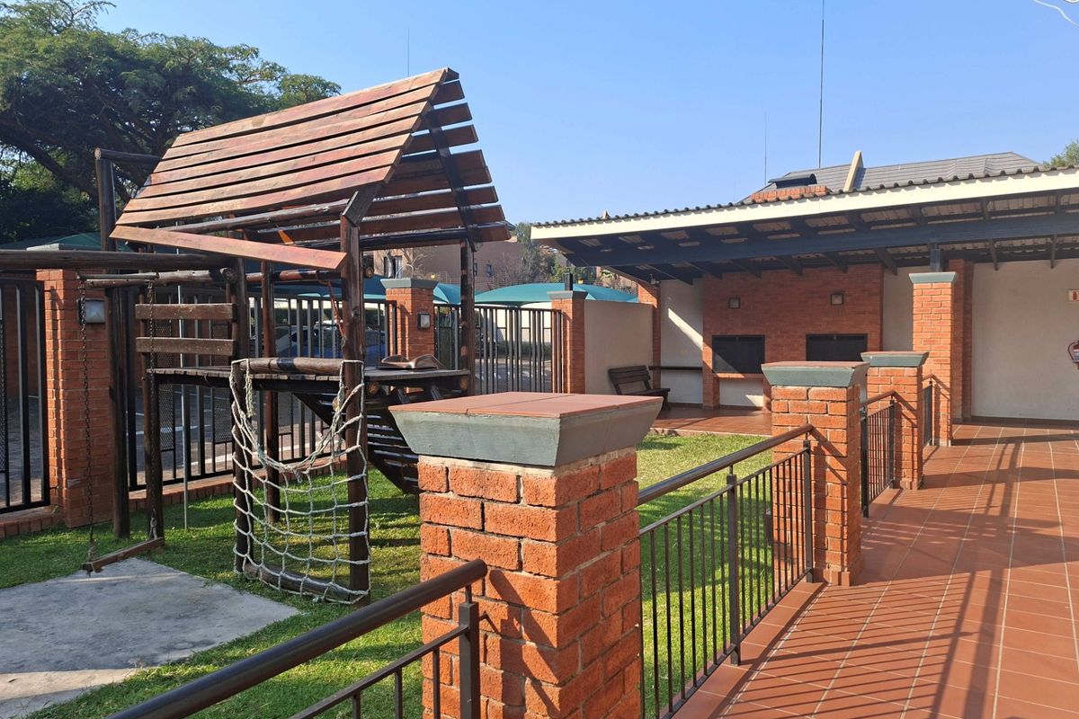 Outdoor communal area featuring a tiled patio, built-in braai facilities, and a wooden playground structure set within a landscaped garden space.