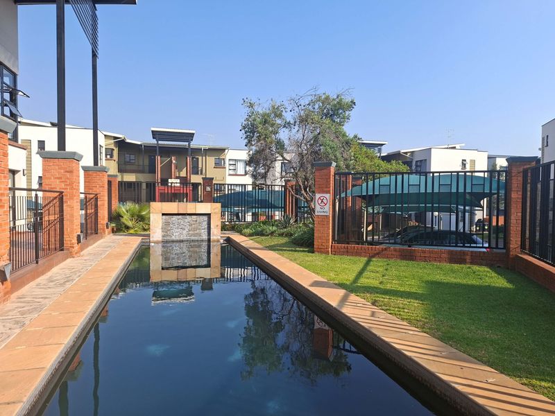 A well-maintained communal swimming pool area featuring a decorative water feature, brick perimeter walls, and manicured lawn within an apartment complex.