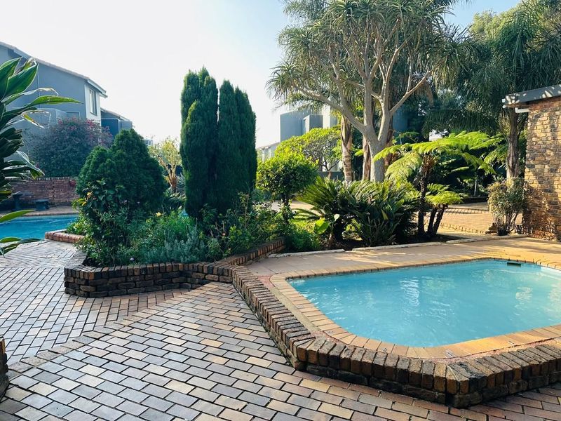 A well-maintained outdoor pool area featuring brick paving, a raised brick-edged swimming pool, and established garden landscaping.