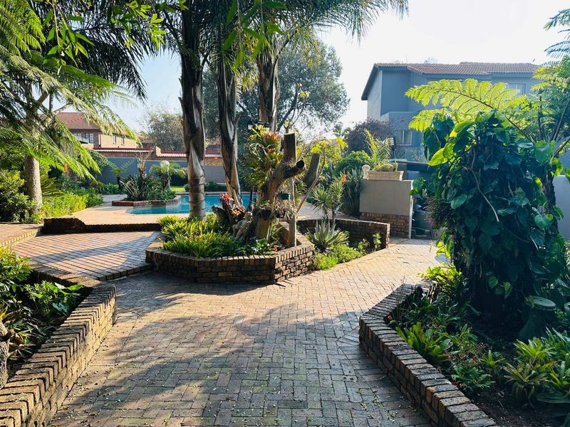 A well-maintained outdoor pool area featuring brick paved pathways, established garden beds, and mature trees surrounding a swimming pool.
