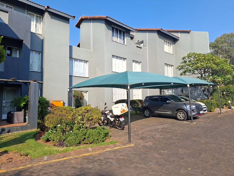 Front exterior view of a multi-story apartment complex featuring a paved driveway with covered shade cloth parking and established garden landscaping.