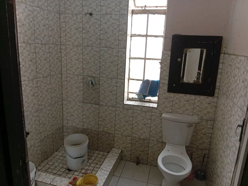 A compact bathroom featuring a tiled shower area, standard toilet, and steel-framed window, requiring renovation to update finishes and fixtures.
