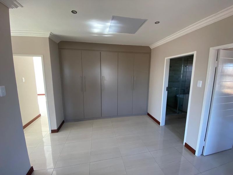 A clean, empty bedroom featuring neutral tiled flooring, modern built-in cupboards, recessed lighting, and direct access to an en-suite bathroom.