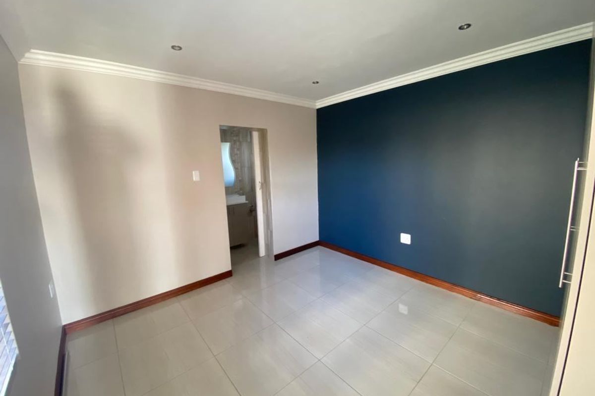 A clean, empty bedroom featuring neutral and dark blue accent walls, tiled flooring, and recessed downlighting, with an adjoining en-suite bathroom.