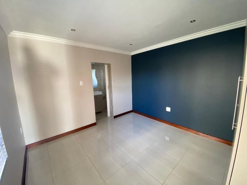A clean, empty bedroom featuring neutral and dark blue accent walls, tiled flooring, and recessed downlighting, with an adjoining en-suite bathroom.